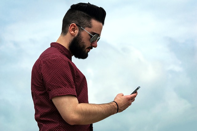 Muž s čiernymi vlasmi a bradou, v kockovanej košeli pozerá do mobilného telefónu